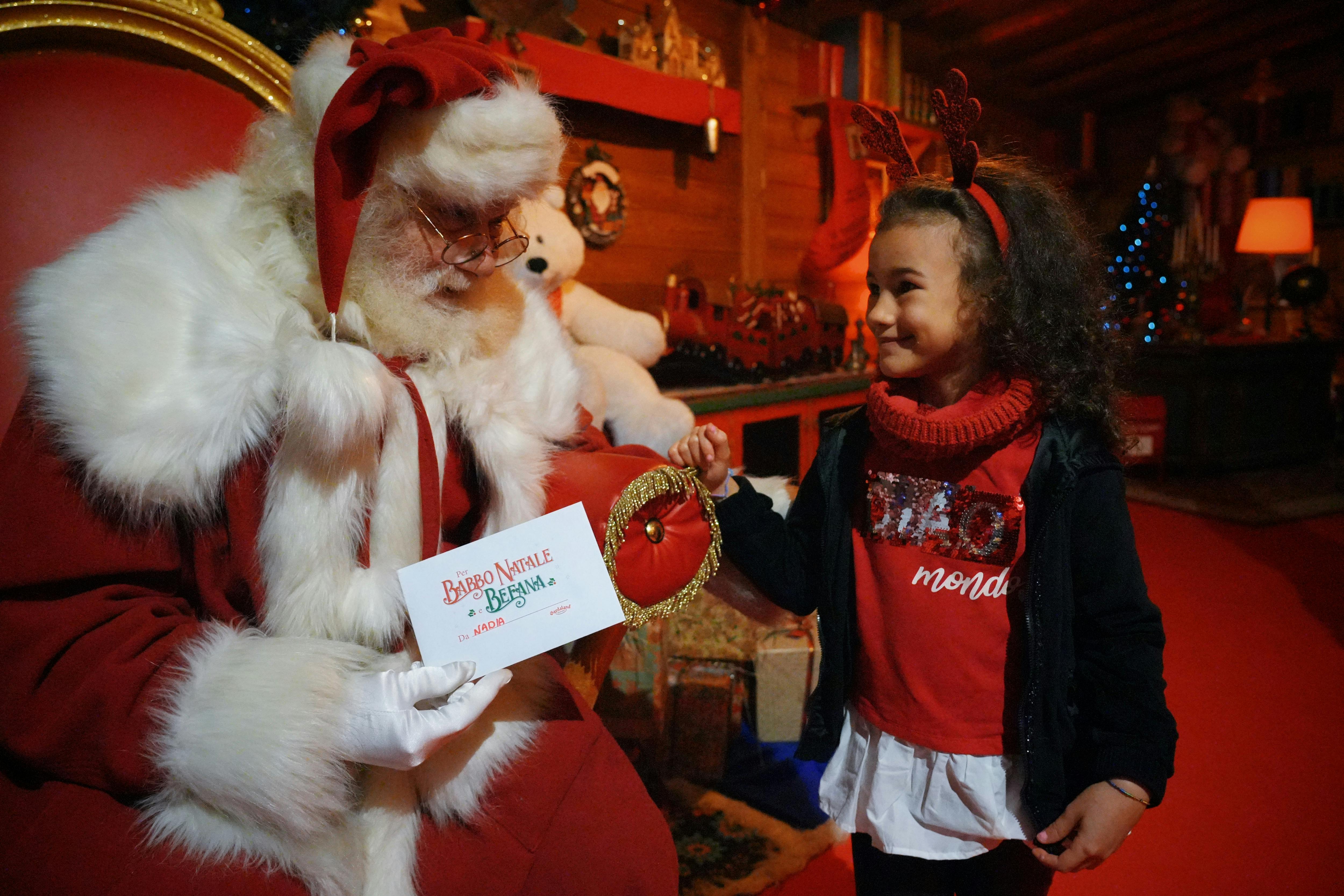 Dítě, které má na sobě sobí parohy a usmívá se na Santa Clause, zatímco dostává červený váček a dopis ve slavnostním krytém prostředí.