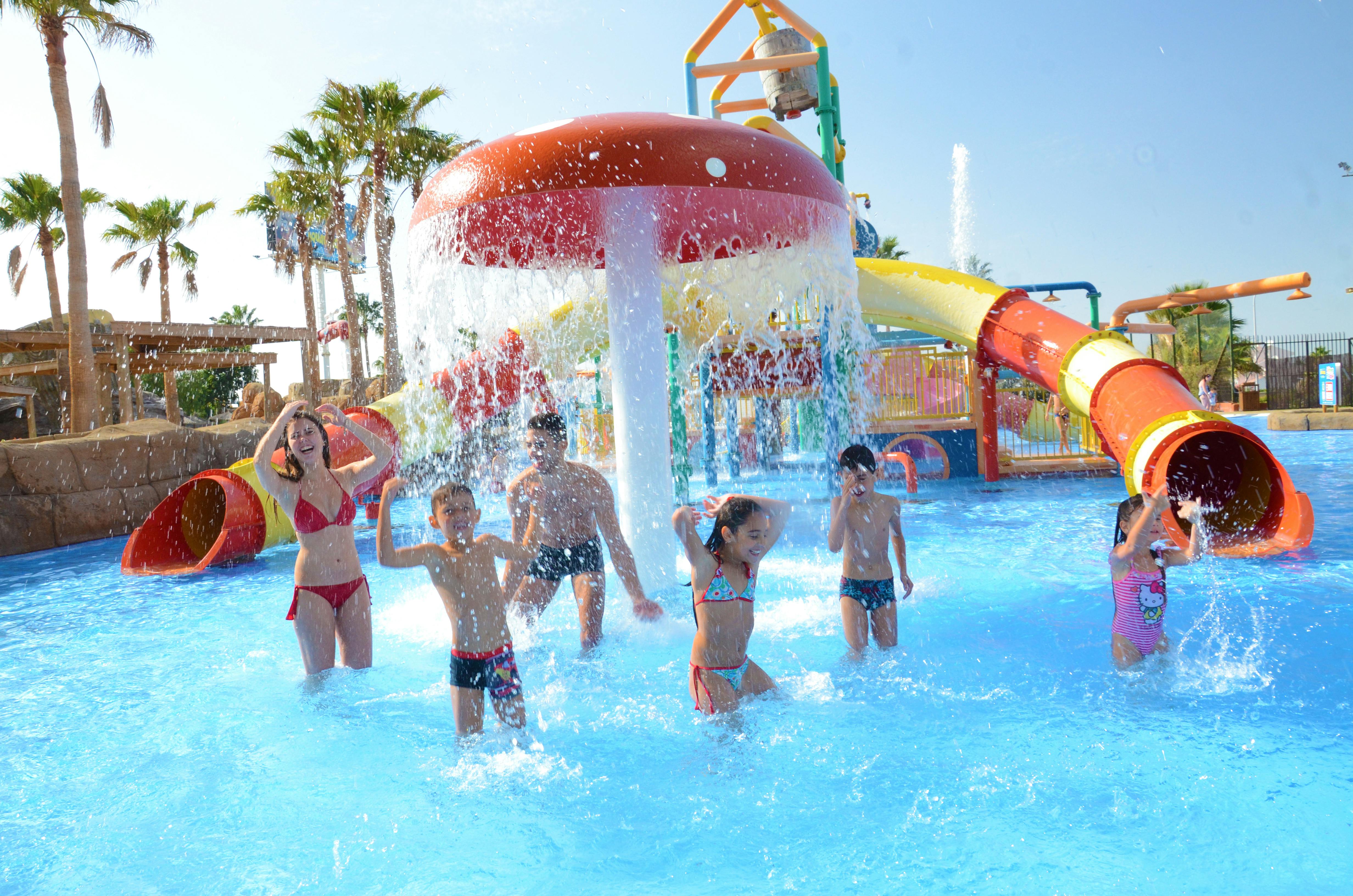Børn og voksne plasker under en stor svampefontæne i et vandland med rutsjebaner og palmer i baggrunden.