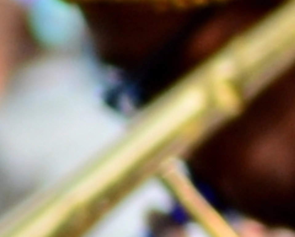 Two musicians perform outdoors; one playing a red trumpet, the other a trombone. A small crowd is visible in the background.
