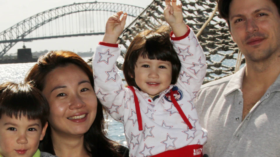 A family of four poses happily on a boat with a large bridge visible in the background.