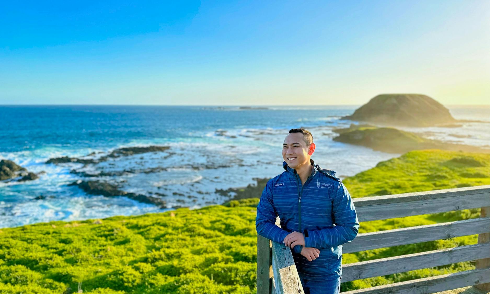 Osoba w niebieskiej kurtce uśmiechnięta, wsparta na drewnianej balustradzie, z malowniczym krajobrazem nadmorskim w tle.
