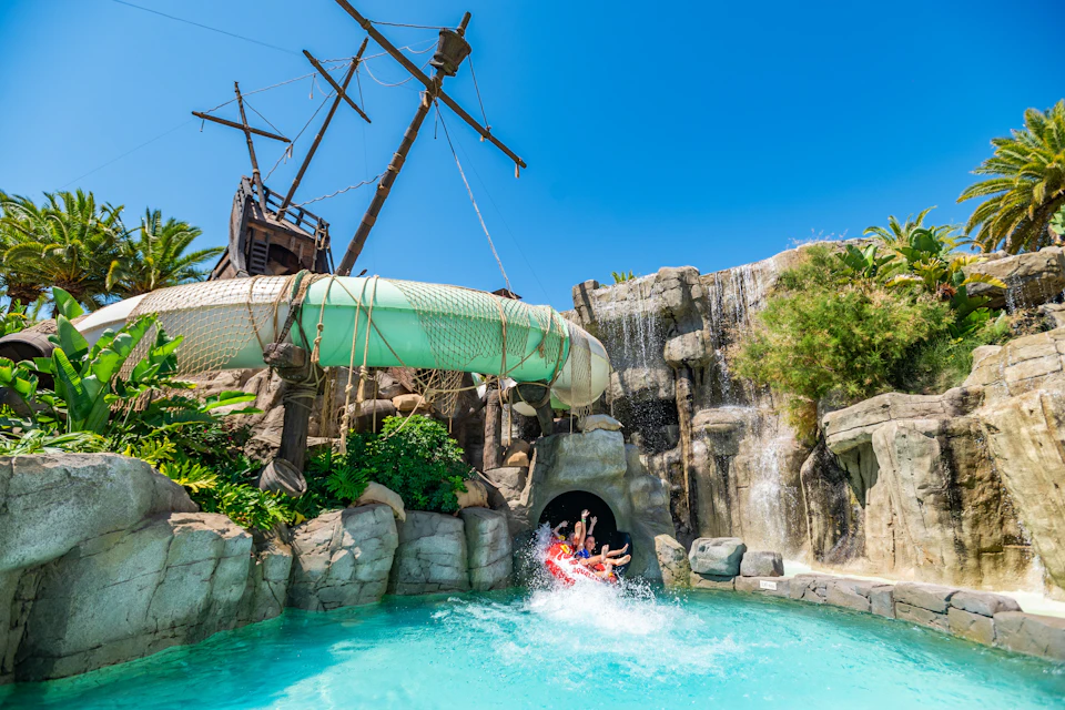 People sliding out of a waterslide into a pool, surrounded by rocks, a waterfall, and a pirate ship structure.