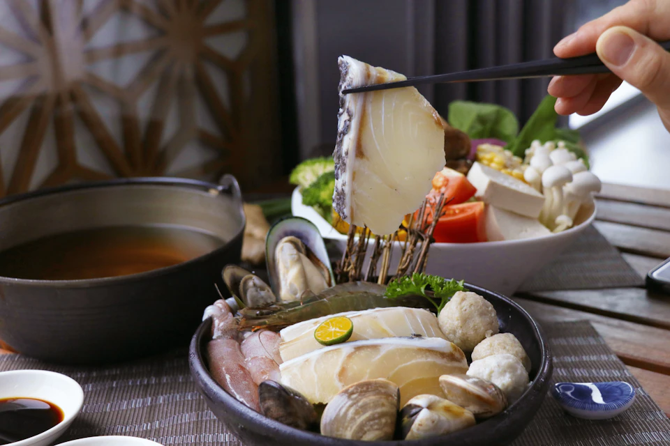 A bowl of assorted seafood and vegetables with a piece of fish being lifted by chopsticks.