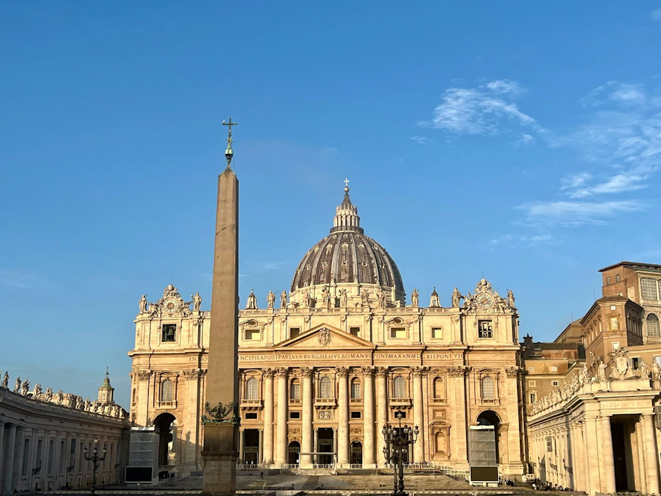 St. Peter's Basilica in Vatican City, featuring its prominent dome and an obelisk in St. Peter's Square under a clear blue sky.