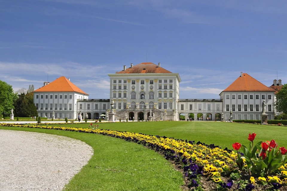 Large white palace with red roofs, ornate architecture, and manicured gardens with yellow and purple flowers under a clear blue sky.