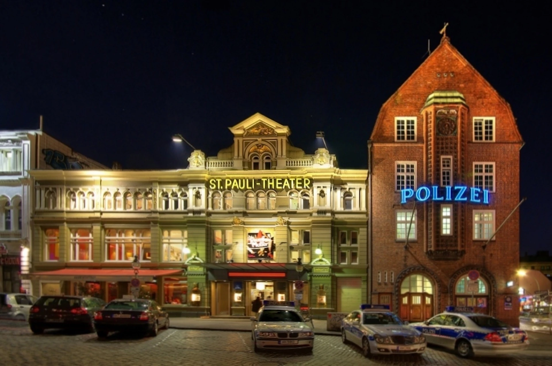 At night, the lit-up St. Pauli Theater facade is next to a building with a blue "Polizei" sign. Several police cars are parked in front.
