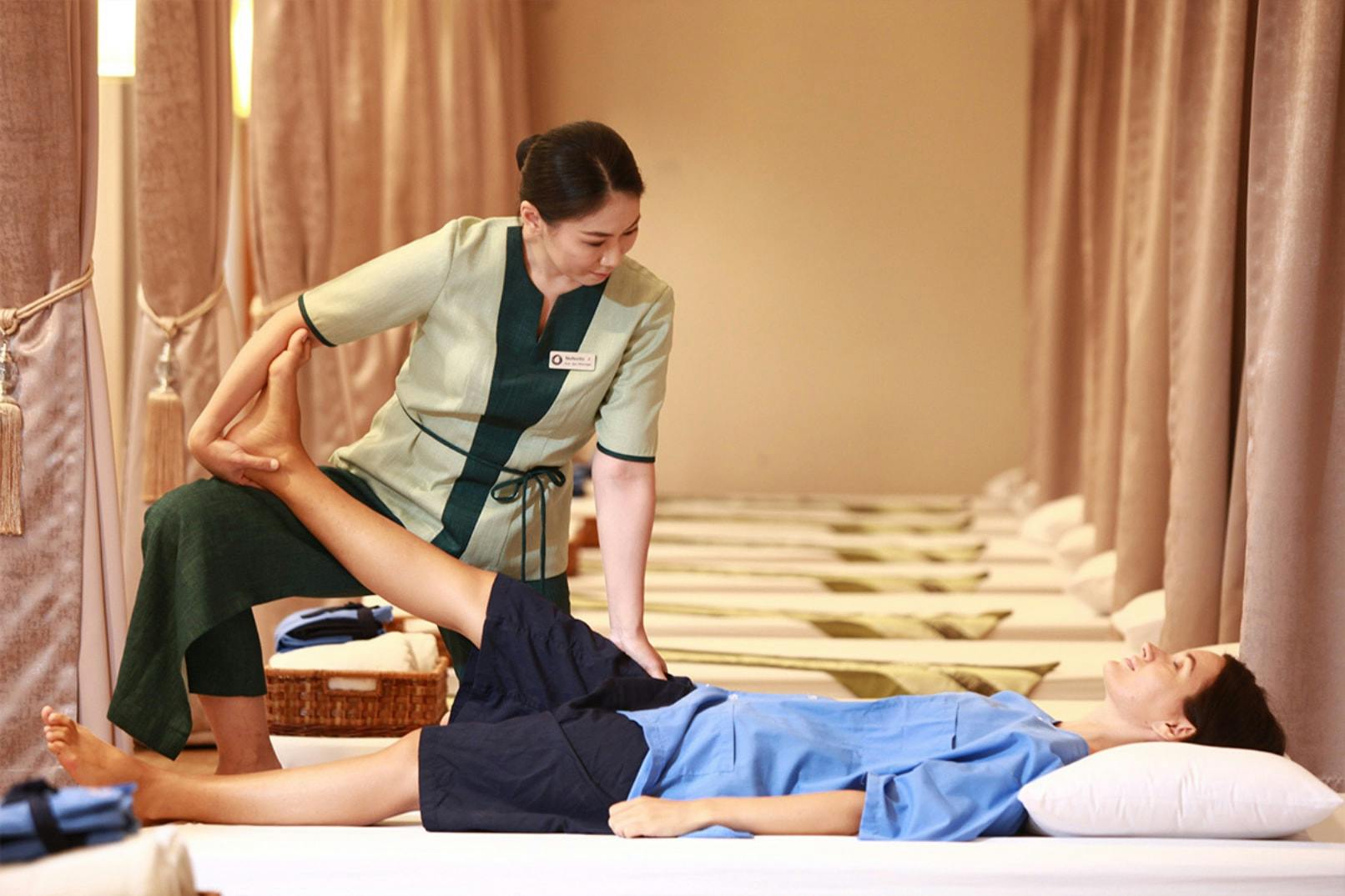 A therapist in green attire assists a person lying on a massage table in a stretching position in a spa setting.