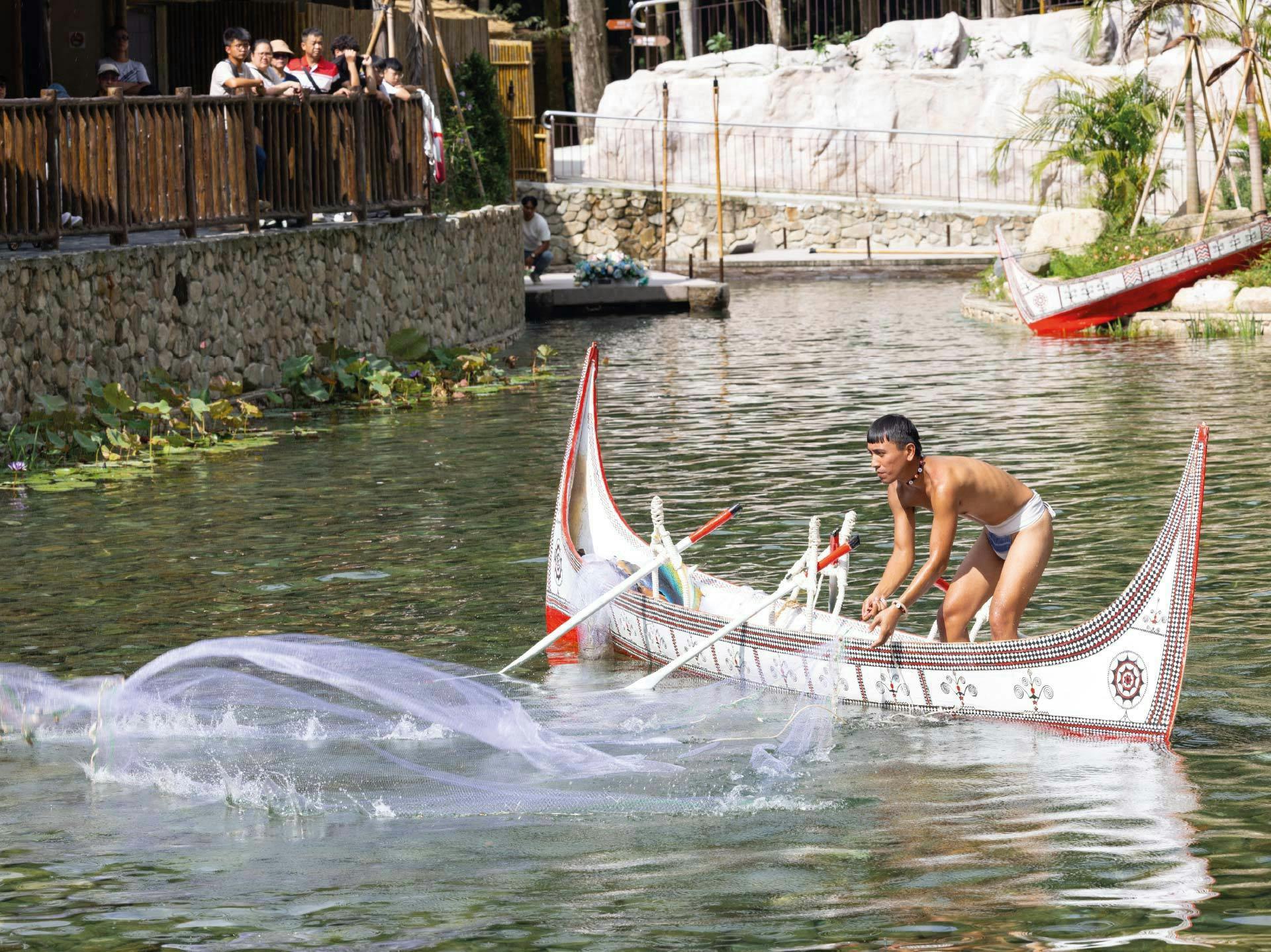 A man in a small, decorated boat on water handling a large fishing net, with a natural backdrop of rocks and plants.