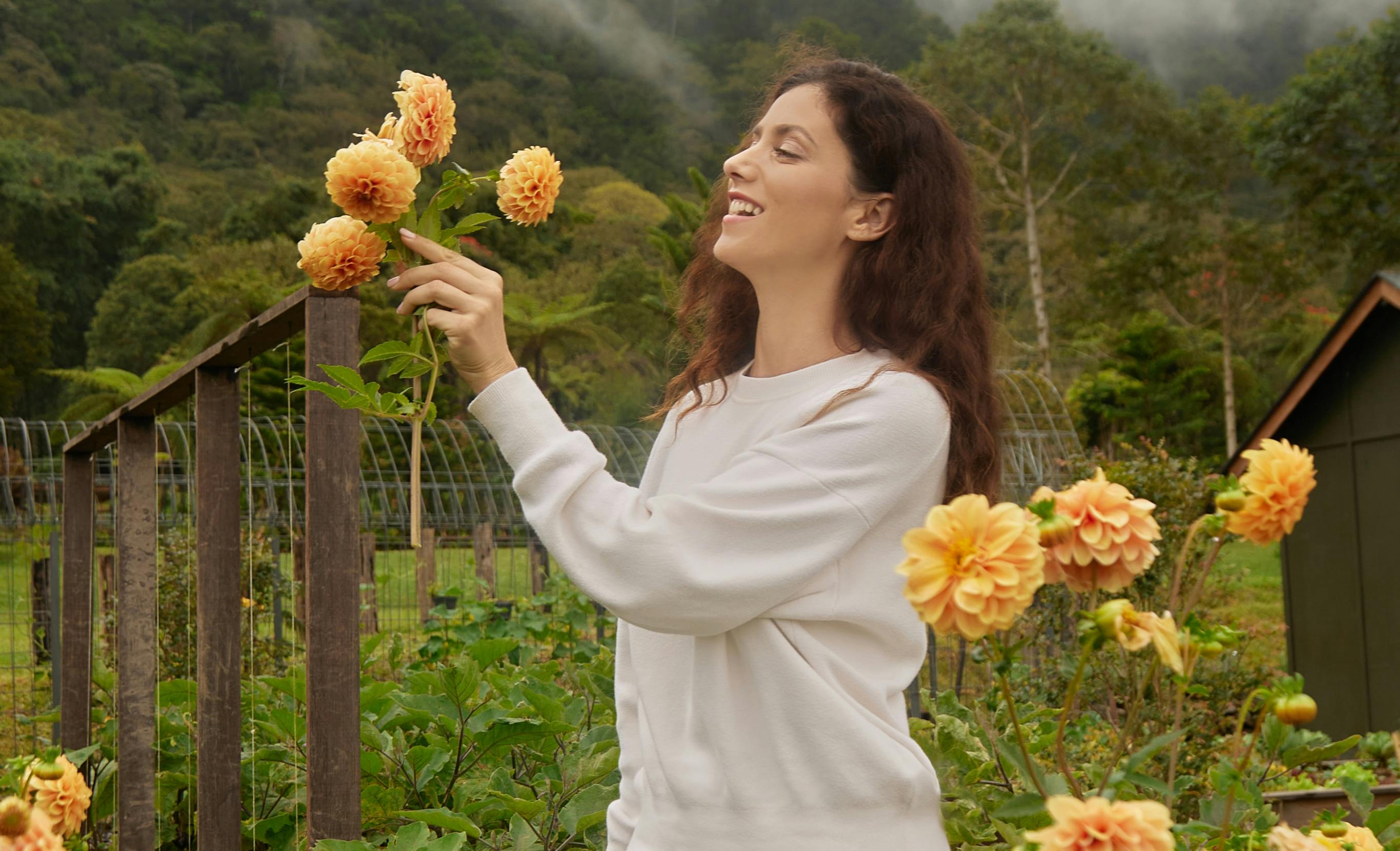 A person in a garden smiling while interacting with orange flowers, with a lush green background and misty hills.