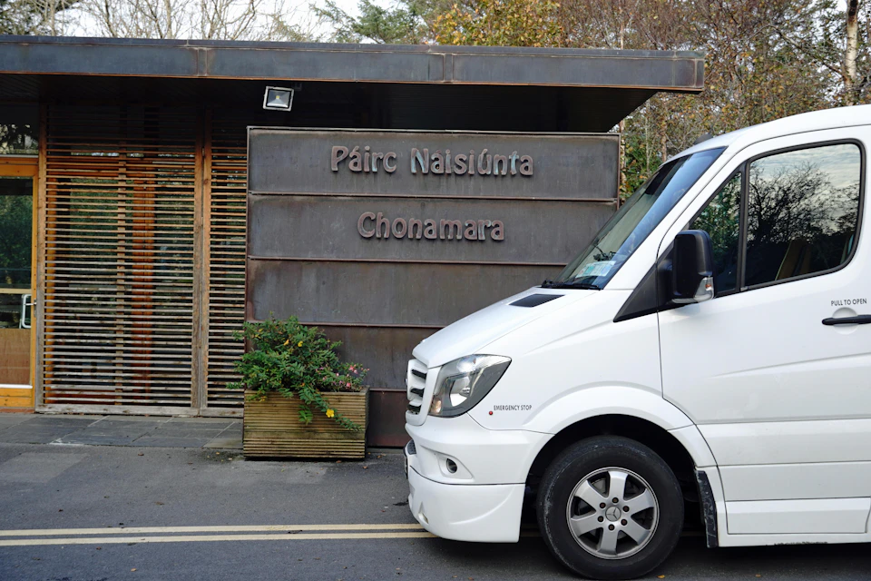 White van parked in front of a building with a sign reading "Páirc Naisiúnta Chonamara" and a planter with green foliage.