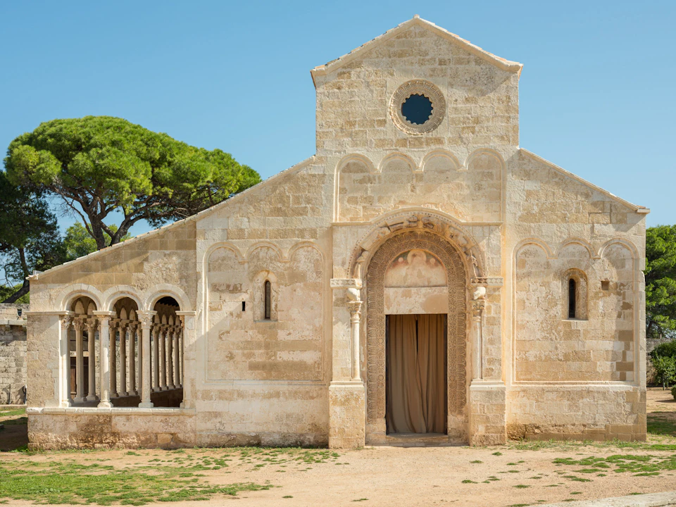 An old stone church with arched windows and a central entrance, flanked by a columned section. Trees are visible in the background.