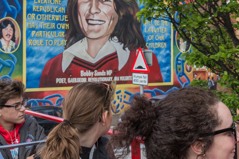 A group of people on an open-top bus look at a large mural depicting a person with text on a building wall.