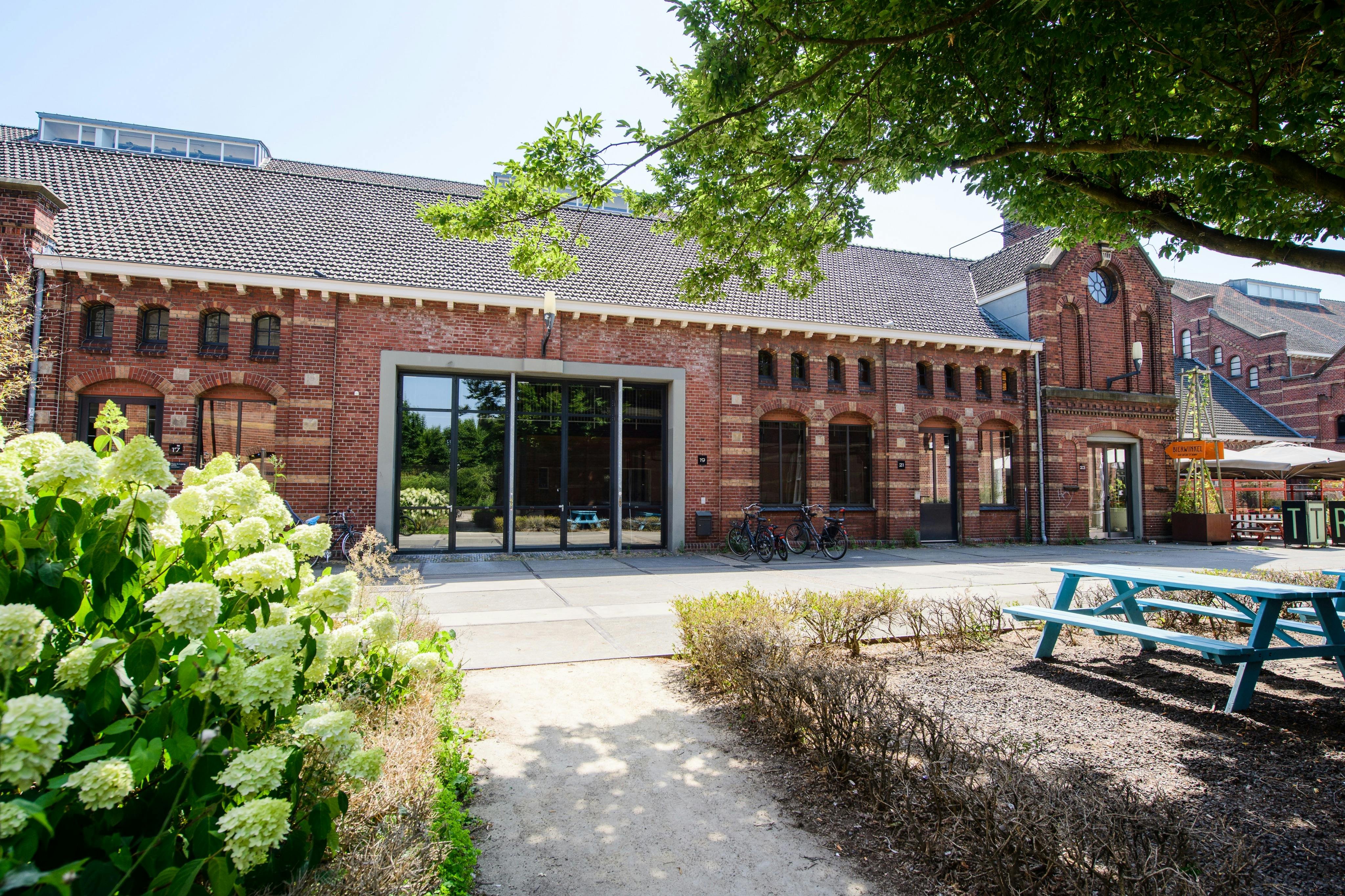 红砖建筑，有大窗户，外面停放着自行车，还有一条通往花卉和树木的花园区的小路。
