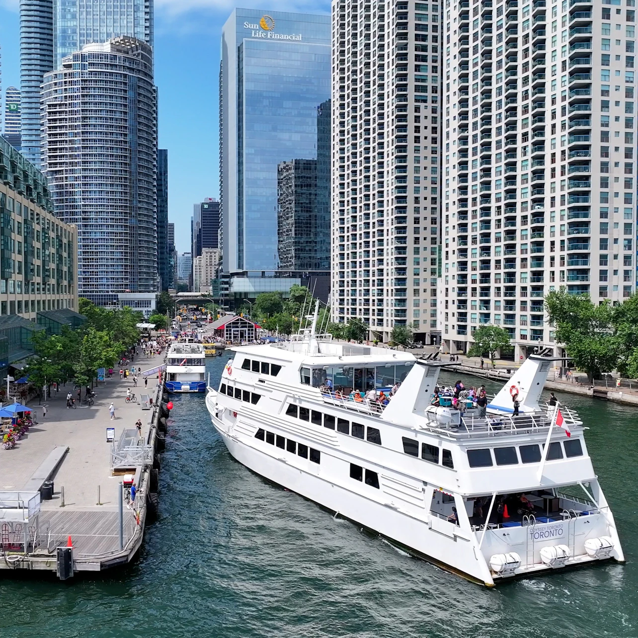 A white sightseeing boat is docked along a river surrounded by tall, modern buildings and a bustling promenade.