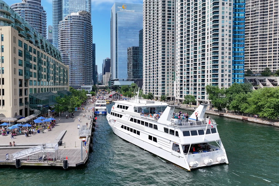 A white sightseeing boat is docked along a river surrounded by tall, modern buildings and a bustling promenade.