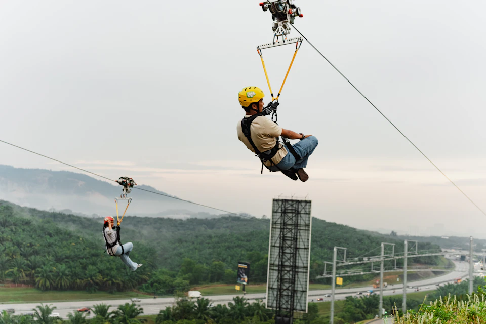 Two people ziplining over a green landscape with roads and hills in the background under a cloudy sky.