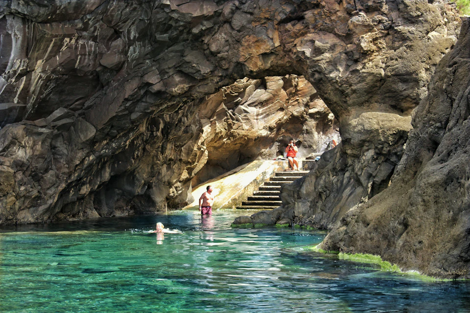Two people swimming in clear turquoise water inside a rocky cave, with another person sitting on stone steps nearby.