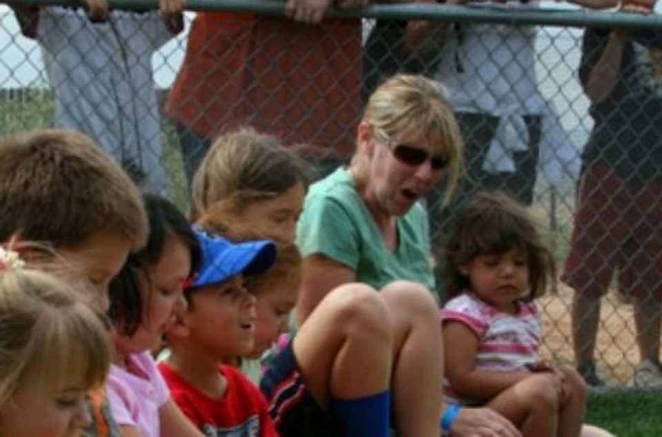 Children sitting on grass, lined up and touching a large yellow snake draped across their laps. Adults and a fence in the background.