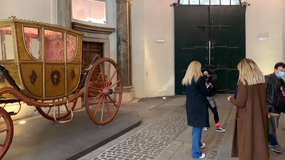 Two women standing near a display of an antique carriage with large wooden wheels in a historic building with cobblestone flooring.