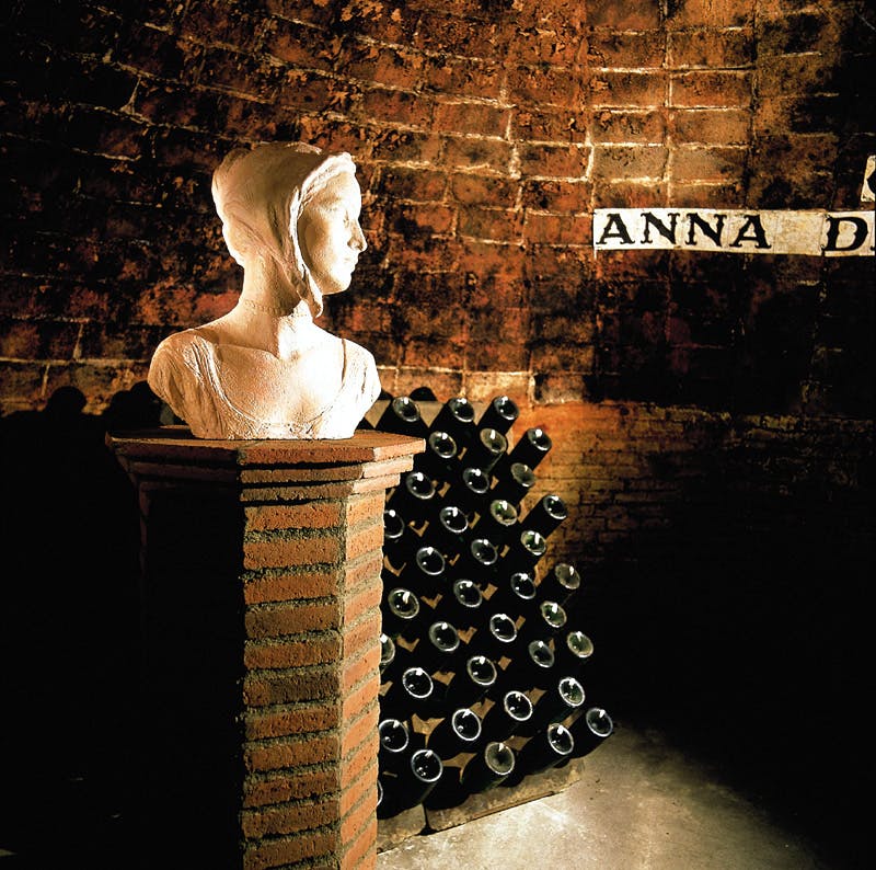A bust of a woman on a brick pedestal in a cellar, with a wall of wine bottles and a partial sign reading "ANNA D".