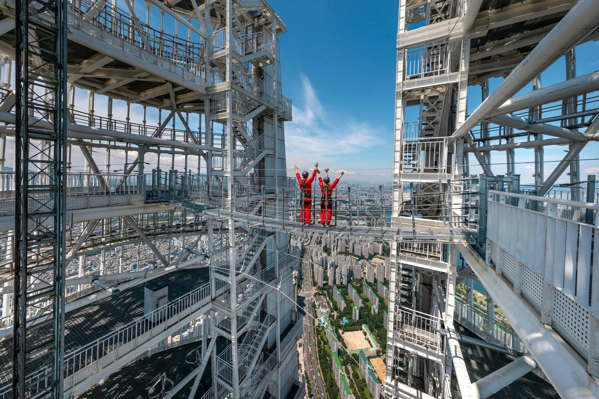 Dos personas vestidas con monos rojos y los brazos levantados están de pie en una pasarela elevada entre estructuras de acero, con vistas a un paisaje urbano.
