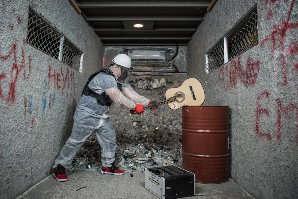 Person in protective gear smashing a guitar on a metal barrel in a narrow, debris-filled alleyway. Broken equipment on the floor.