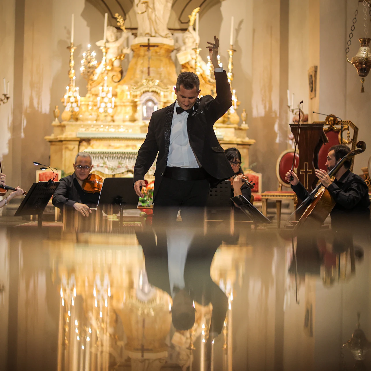 A man in a suit gestures dramatically in front of an orchestra inside a grand, ornate church, with his reflection visible below.
