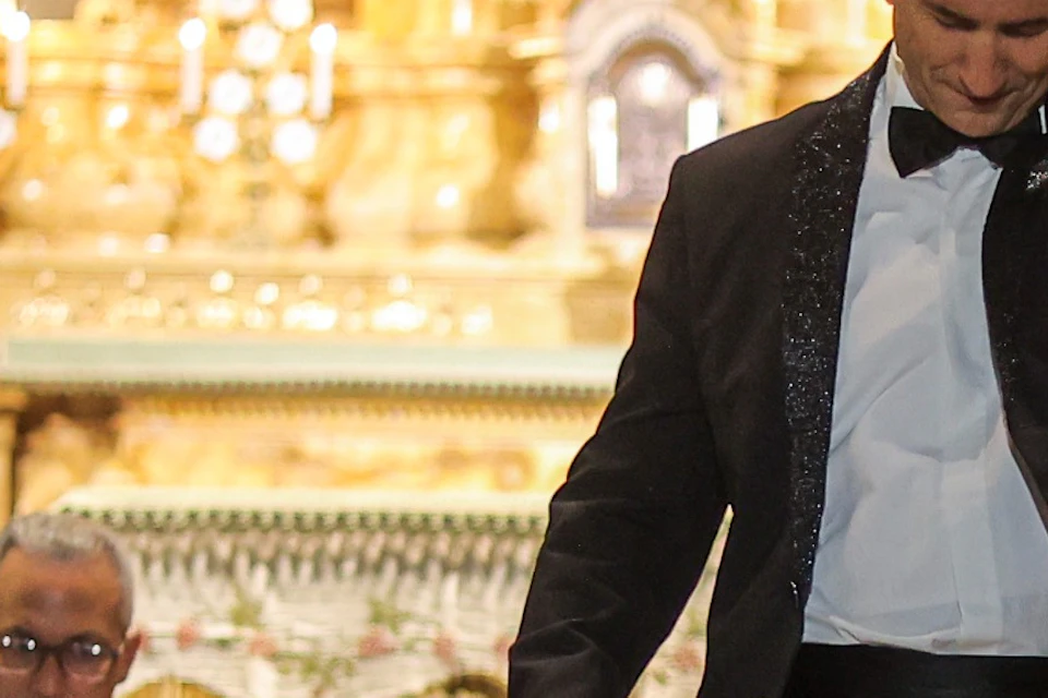 A man in a suit gestures dramatically in front of an orchestra inside a grand, ornate church, with his reflection visible below.