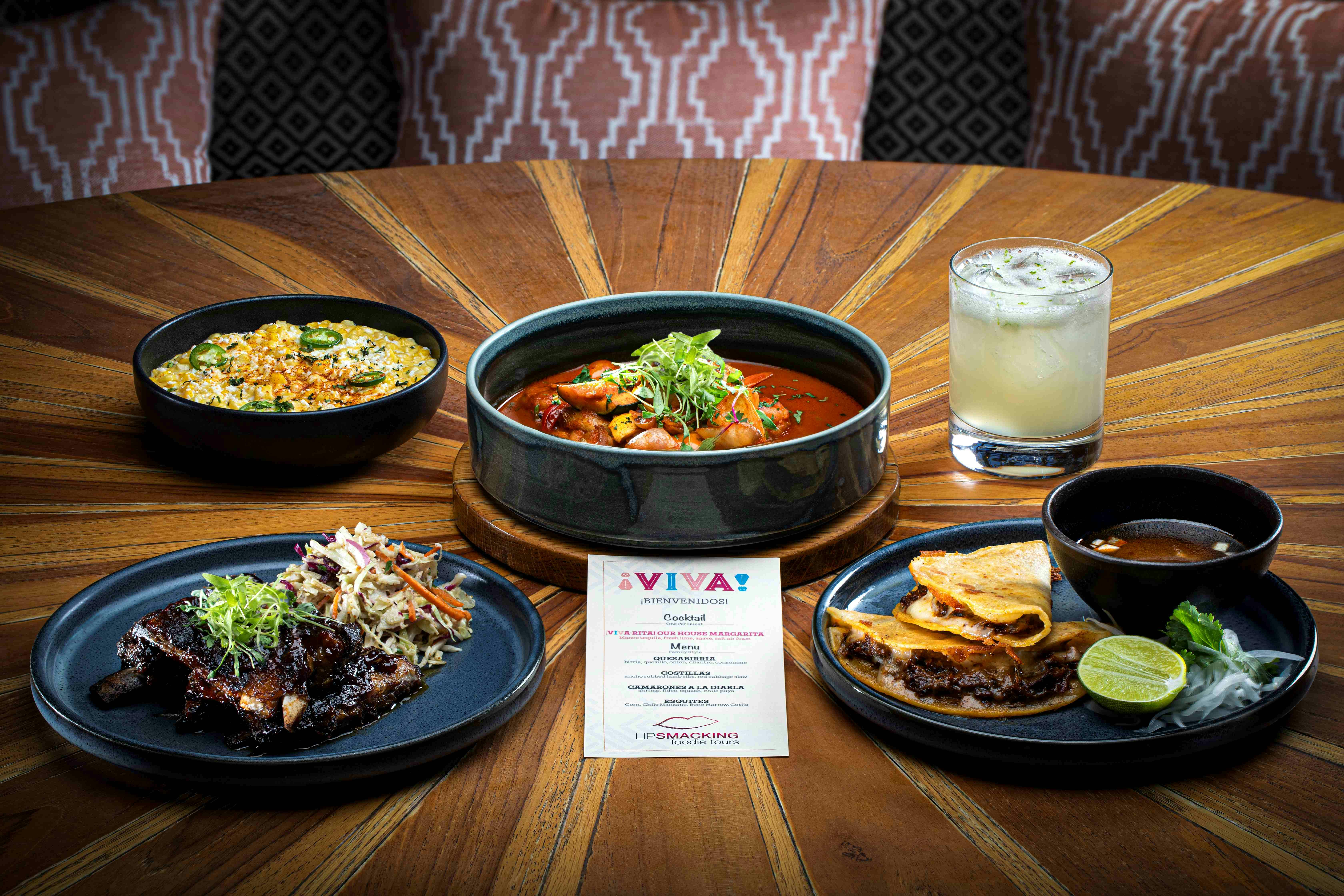 Ένα ξύλινο σετ τραπεζιού με διάφορα πιάτα, όπως κάρυ, τάκος, ρύζι και ένα ποτό, με μια κάρτα μενού στο κέντρο.