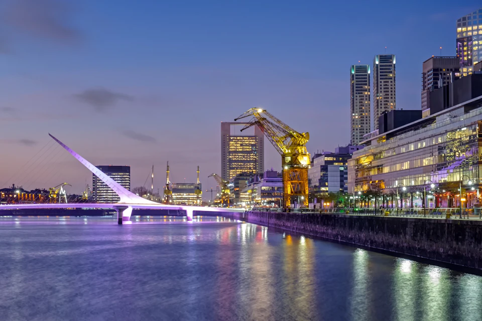 Urban waterfront scene at dusk with a modern bridge, illuminated crane, and city buildings reflecting in the water.