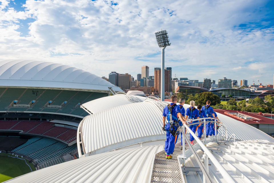 People in blue jumpsuits walk on a rooftop platform above a large stadium, with a city skyline visible in the background.