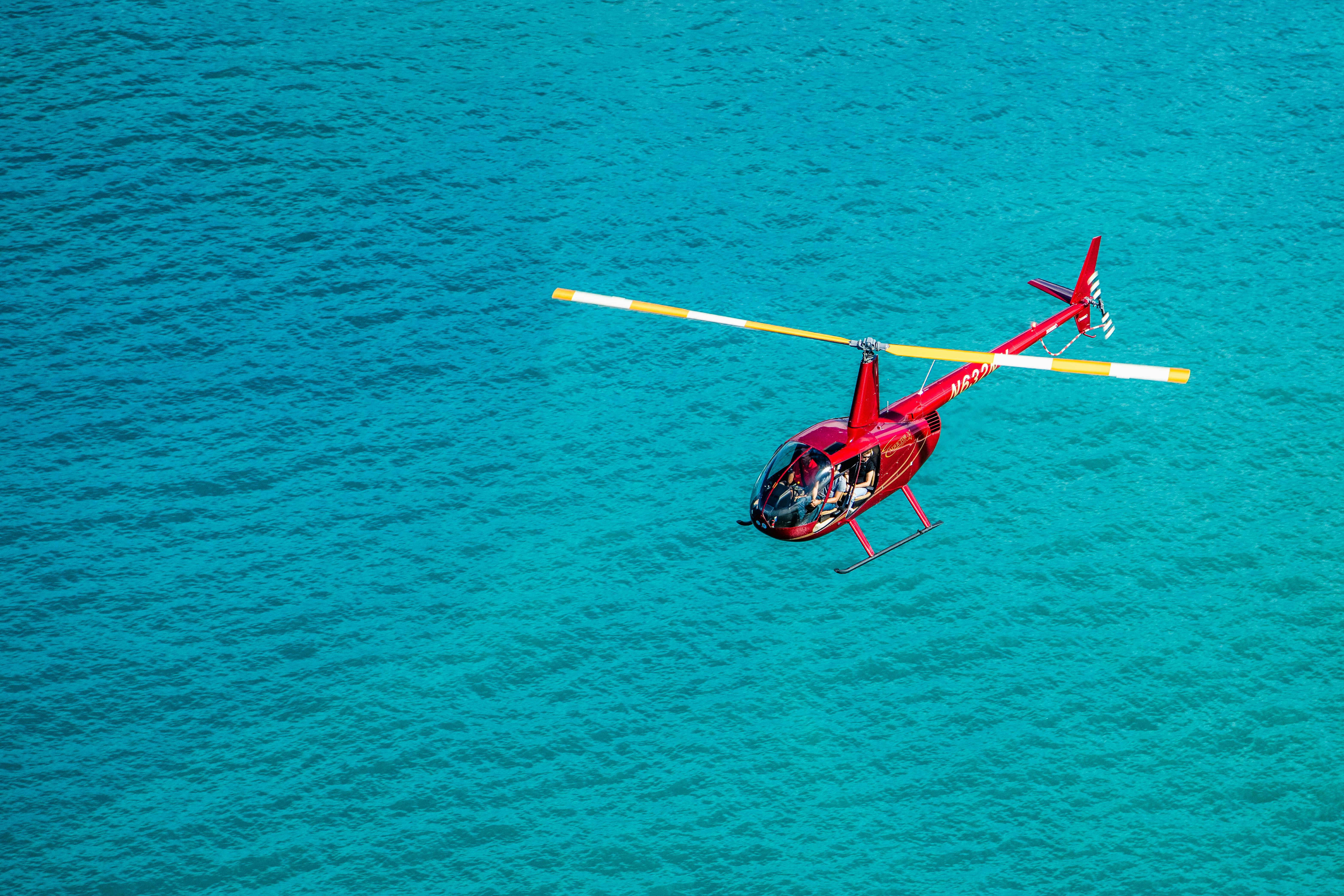 A red helicopter with yellow-tipped rotor blades flies over clear, turquoise water.