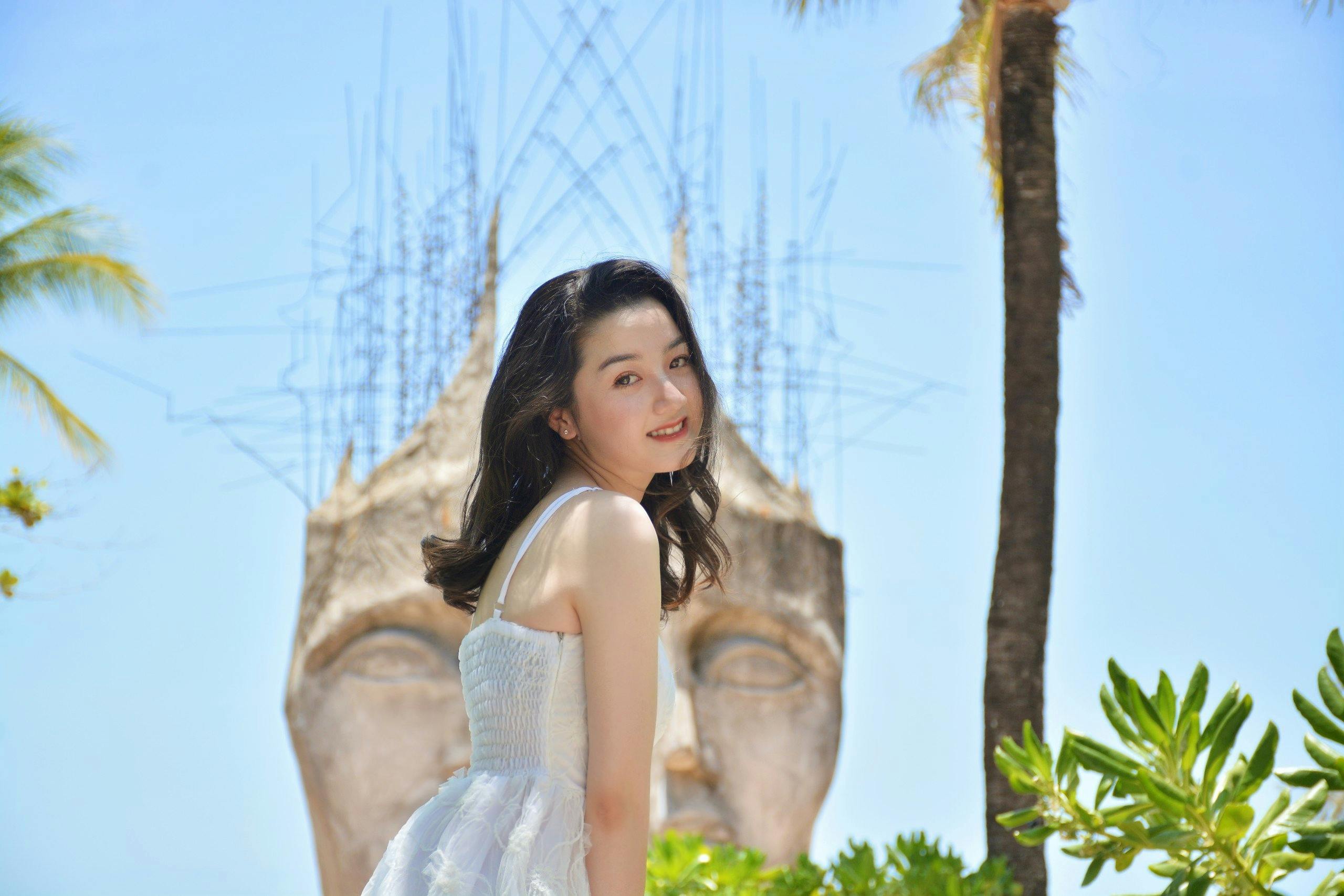 A woman in a white dress smiles outdoors, with a large abstract sculpture of a face and trees in the background.