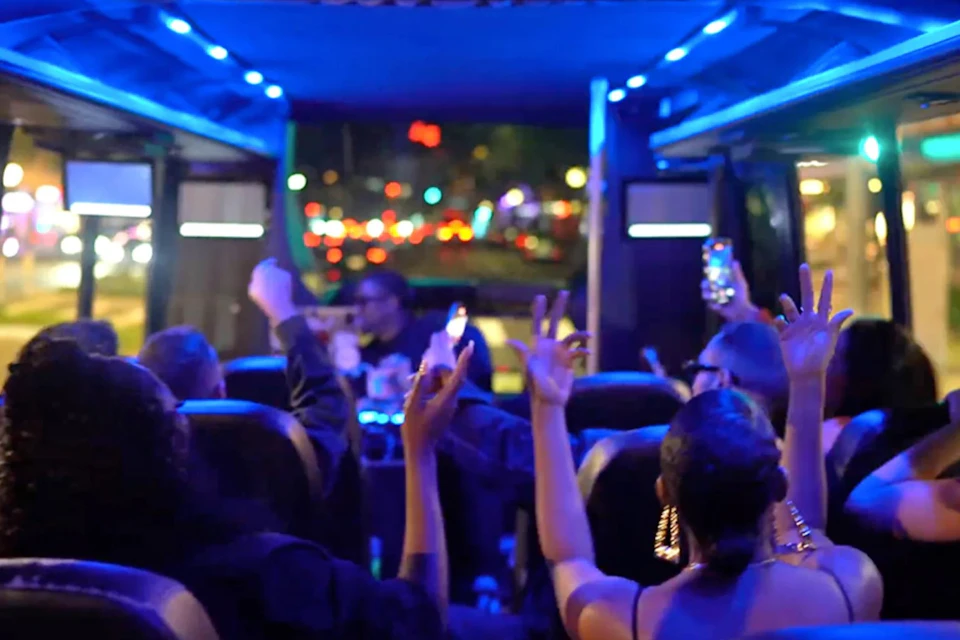 People inside a party bus at night, raising their hands and taking photos, with colorful lights and a blurred cityscape outside.