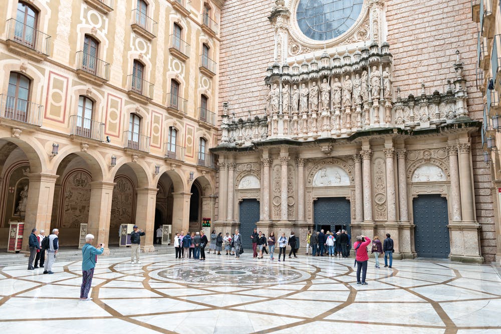 People gather and take photos in a large courtyard with ornate architectural details and a circular mosaic floor design.