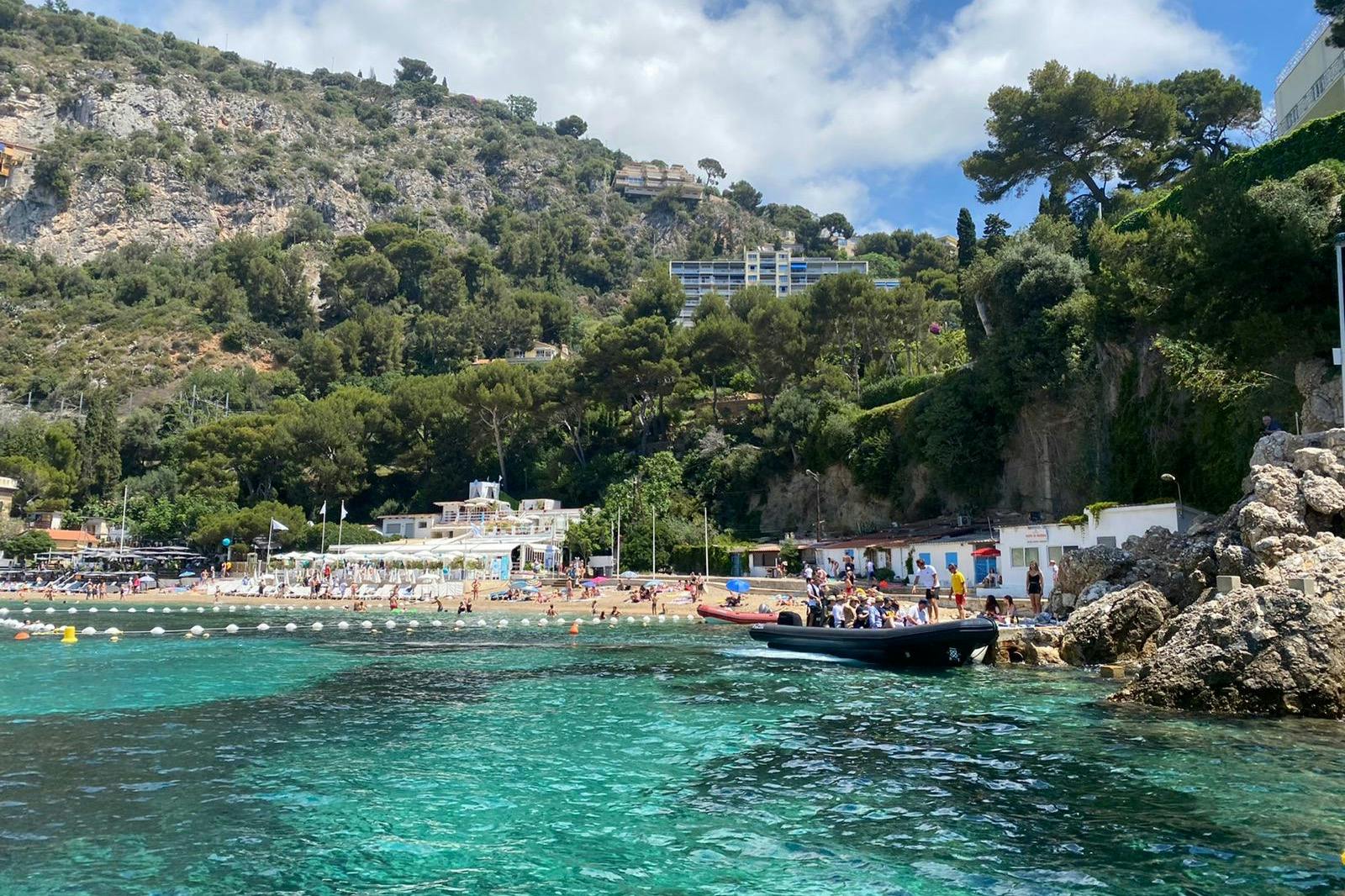 在部分多云的天空下，海滩、绿色山丘、建筑物和停靠在绿松石水上的船只组成的海岸景观。