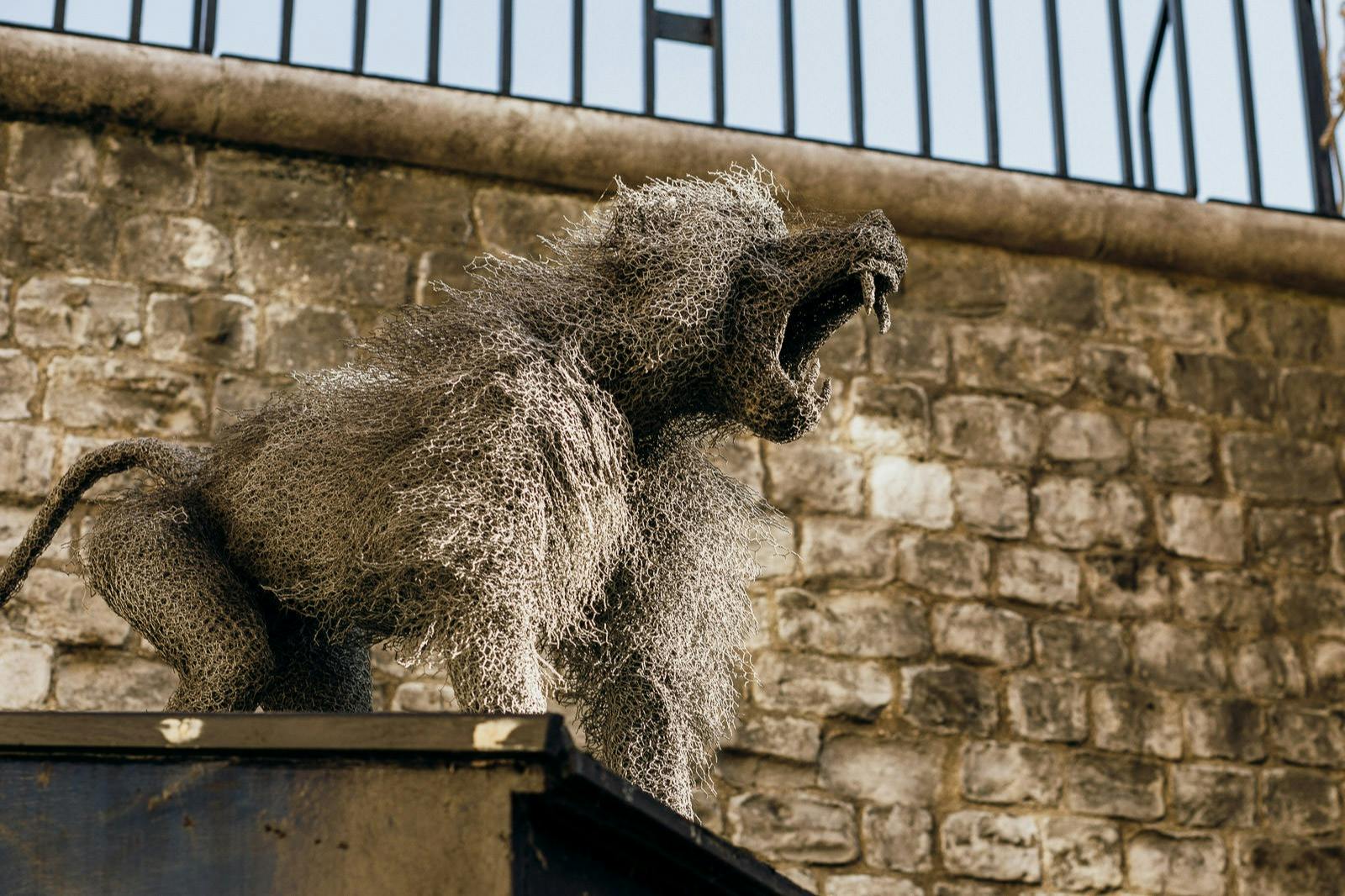 A wire mesh sculpture of a roaring lion stands against a brick wall and a metal fence.
