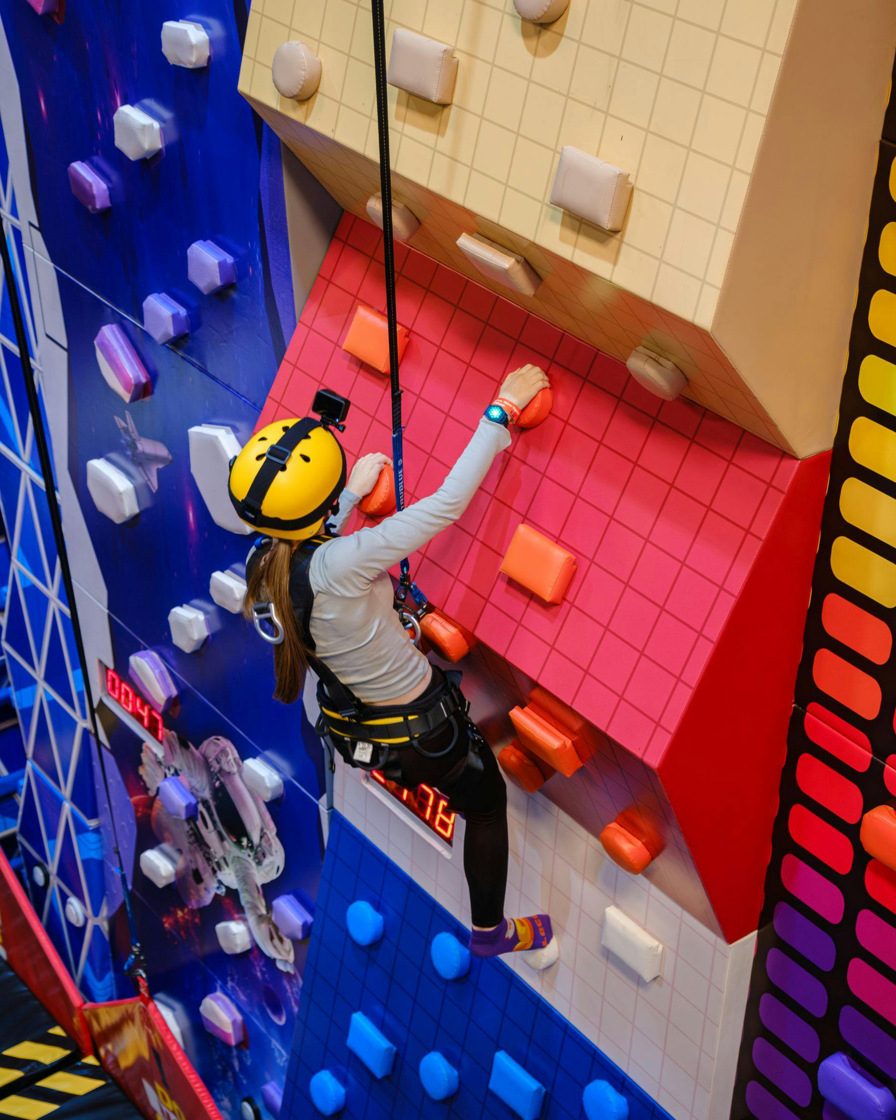 Ένα άτομο που σκαρφαλώνει σε έναν πολύχρωμο τοίχο αναρρίχησης εσωτερικού χώρου φορώντας εξοπλισμό ασφαλείας, συμπεριλαμβανομένου κράνους και ιμάντα.