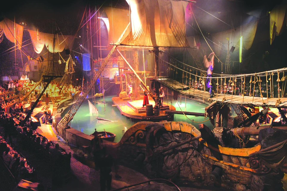 A theatrical pirate ship scene with actors in costume, ropes, sails, water, and stage lights creating a dramatic atmosphere.