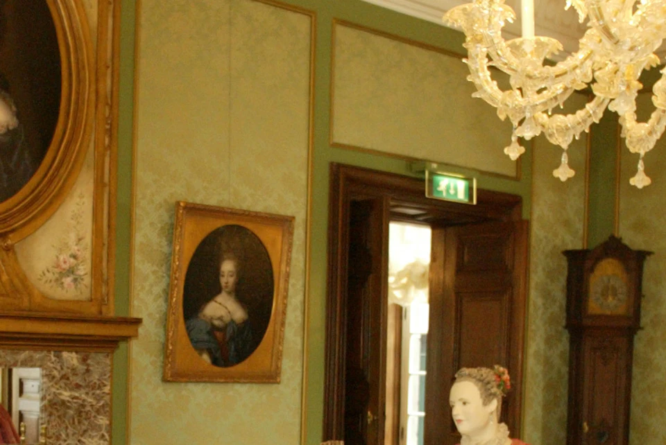A historic room with a chandelier, harpsichord, paintings, and mannequins dressed in 18th-century attire.