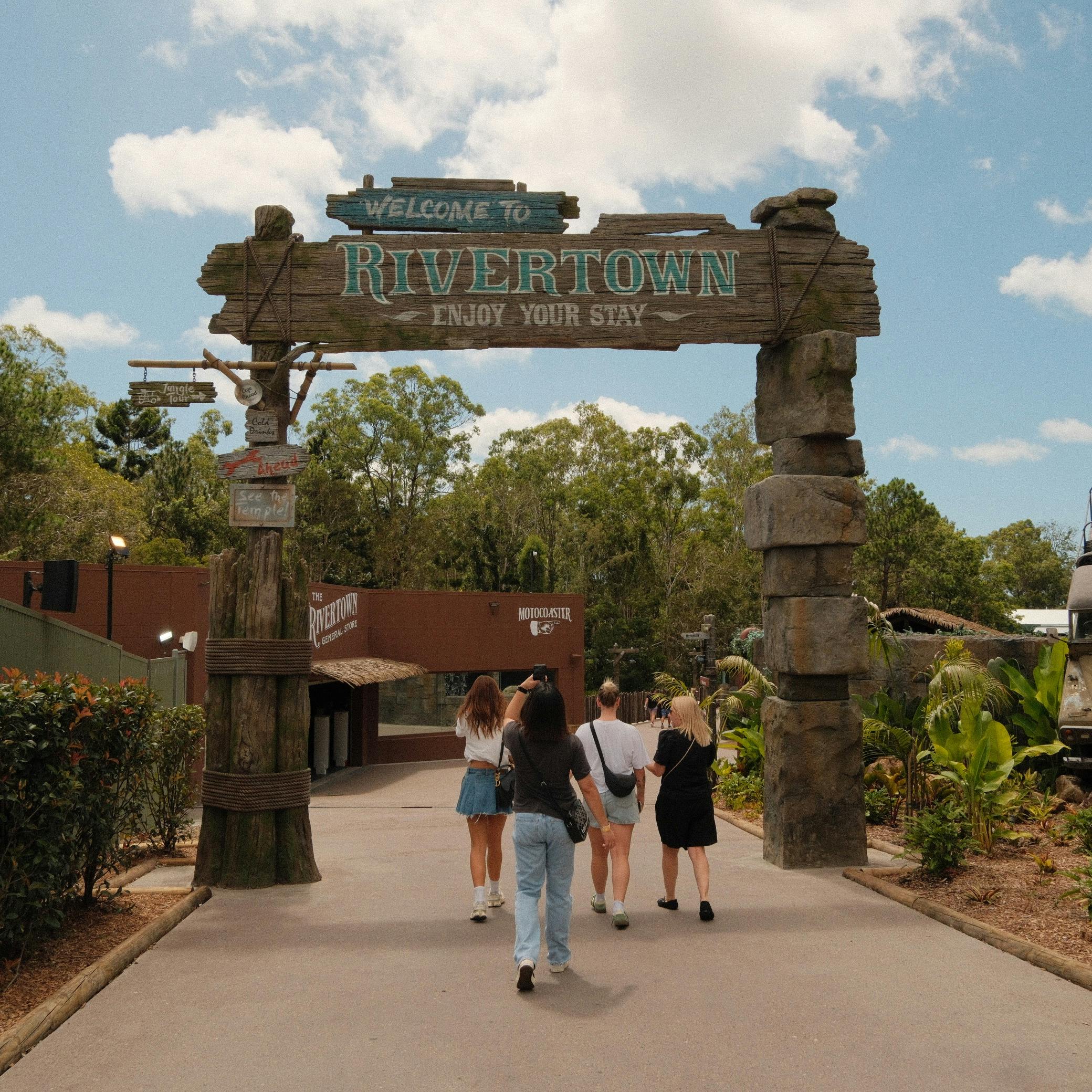 Four people walking under a wooden archway labeled "Welcome to Rivertown, Enjoy Your Stay," surrounded by greenery.