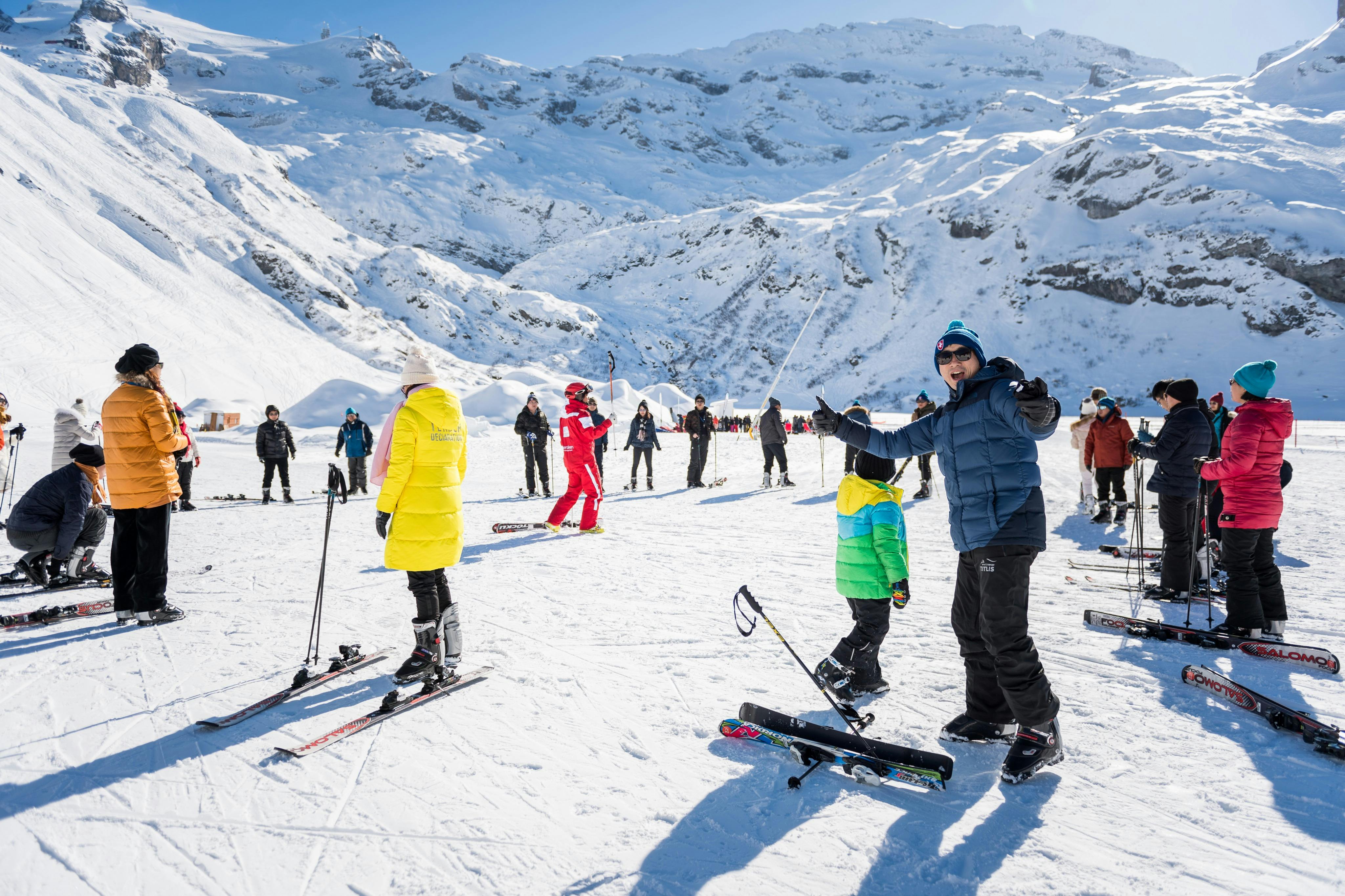 People in colorful jackets and ski gear standing on a snowy mountain slope, with ski equipment scattered around.