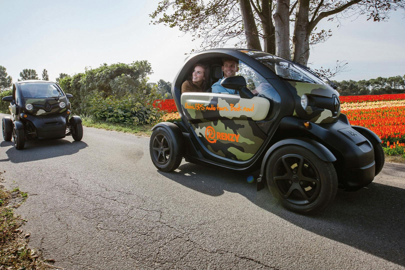 Two people smiling inside a small, camouflaged electric car on a road, with a second similar car behind them. Trees and flowers in the background.