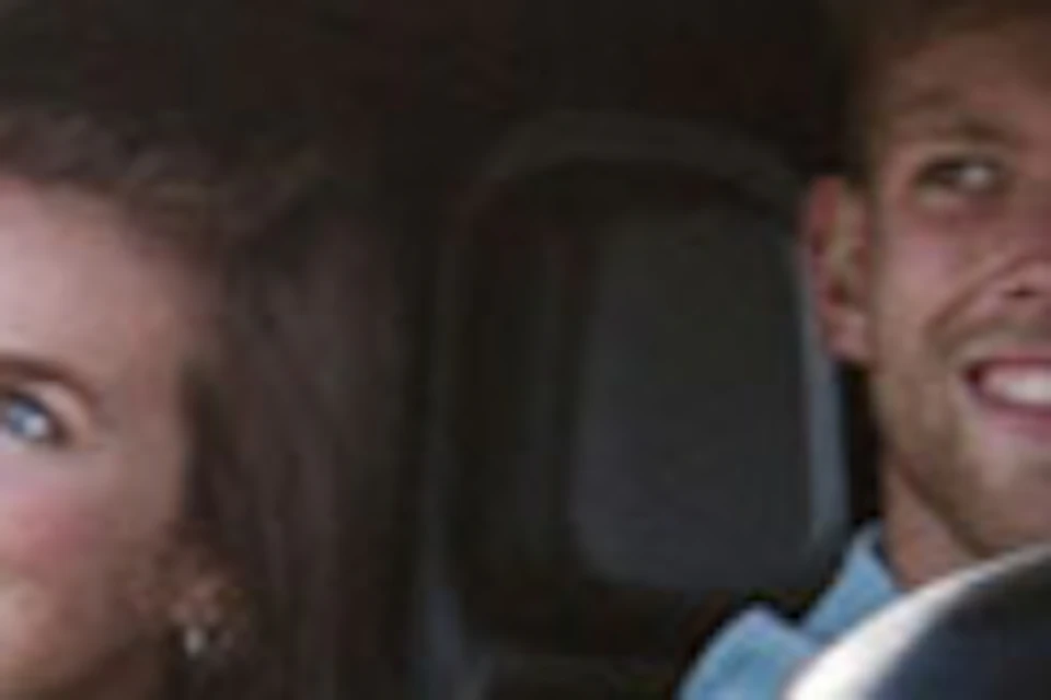 Two people smiling inside a small, camouflaged electric car on a road, with a second similar car behind them. Trees and flowers in the background.
