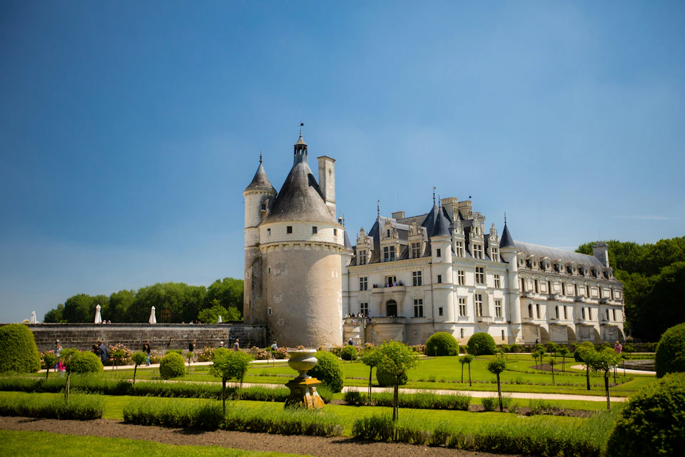 A historic castle with a round tower and a landscaped garden under a clear blue sky.