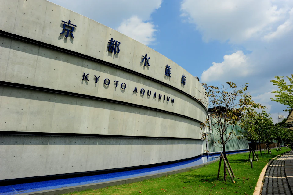 Curved concrete building labeled "Kyoto Aquarium" in English and Japanese, set against a partly cloudy sky with a lawn in front.
