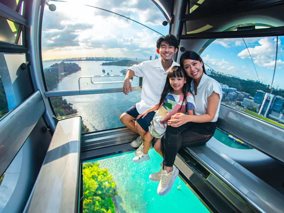 A family of three smiling and seated inside a gondola with a scenic view of water and cityscape visible through the windows.