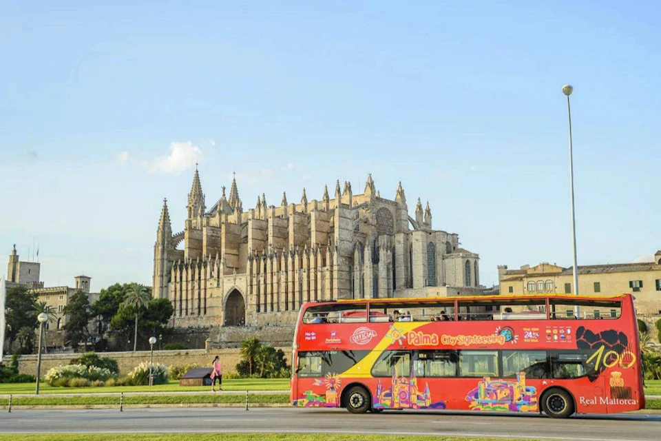 A historic cathedral with intricate gothic architecture stands behind a red double-decker tour bus on a sunny day.