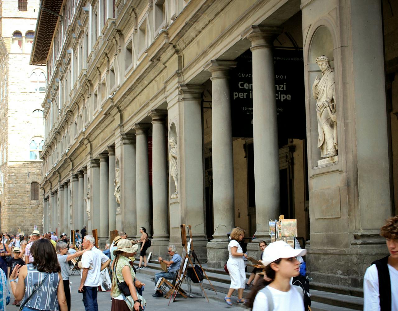 People walk near a historic building with tall columns and classical statues set into alcoves. Art easels and chairs are seen outside.