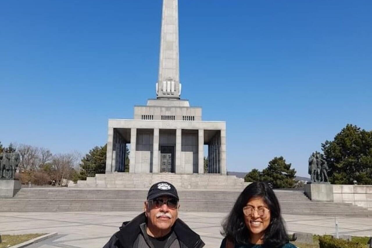 两个人站在一座高高的石碑前，背景是一片晴朗的蓝天。